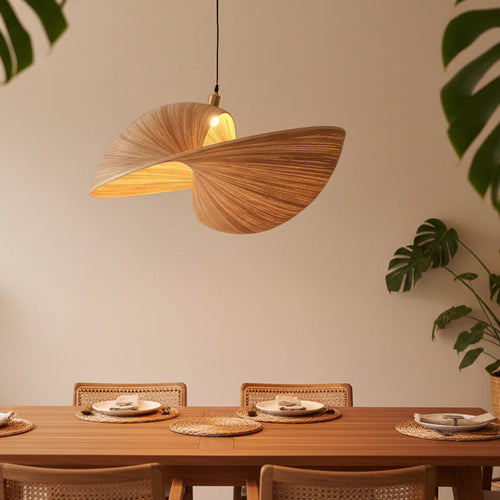 A modern pendant light with a sculptural, shell-like shade made of natural wood grain, glowing with a warm light. It hangs above a wooden dining table set with woven placemats and chairs in a softly lit room with green plants.