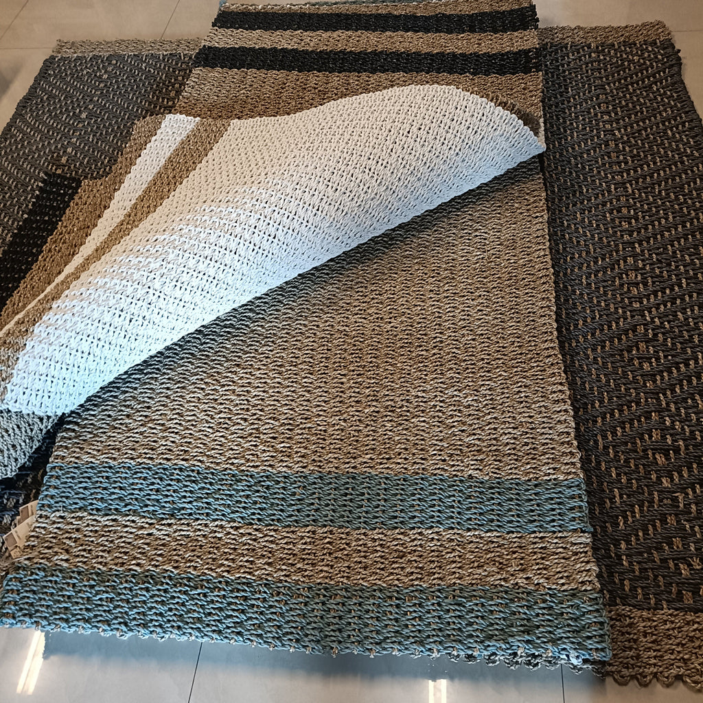 A woven rectangular rug laid on a light tile floor, with one corner folded back to show the underside. The rug has a striped pattern in natural beige, black, and muted blue-gray tones.