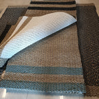 A woven rectangular rug laid on a light tile floor, with one corner folded back to show the underside. The rug has a striped pattern in natural beige, black, and muted blue-gray tones.