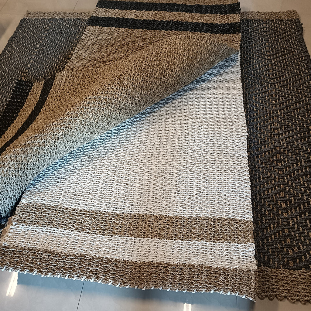 A rectangular woven rug laid on a light tile floor, shown partially folded to display its texture and pattern. The rug features a striped design with bands of black, tan, cream, and light blue-gray.