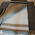 A rectangular woven rug laid on a light tile floor, shown partially folded to display its texture and pattern. The rug features a striped design with bands of black, tan, cream, and light blue-gray.