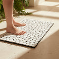 A white pebble bath mat with a black border on a beige tiled floor, with a person's bare feet stepping onto it in warm sunlight next to green plants.