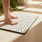 A white pebble bath mat with a black border on a beige tiled floor, with a person's bare feet stepping onto it in warm sunlight next to green plants.