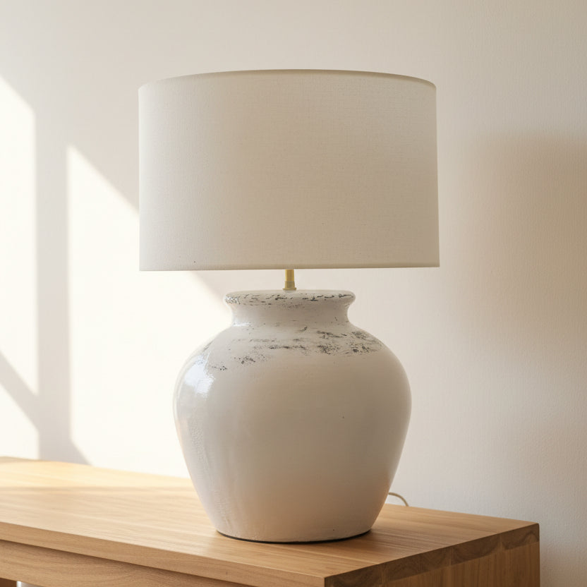 A table lamp with a round glossy off-white ceramic base and a matching cylindrical drum shade, shown sitting on a light wood surface against a warm off-white wall.