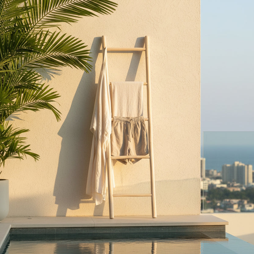 A light natural wood ladder towel rack leaning against a beige stucco wall beside a pool, with a white robe and beige shorts hanging on it. Palm leaves enter from the left, and a city skyline with the sea is visible in the background.