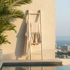 A light natural wood ladder towel rack leaning against a beige stucco wall beside a pool, with a white robe and beige shorts hanging on it. Palm leaves enter from the left, and a city skyline with the sea is visible in the background.