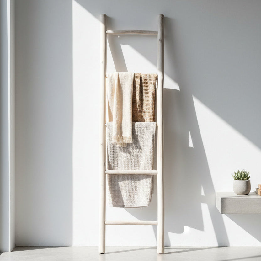 A tall, minimalist wooden ladder towel rack with three rungs, styled with three textured beige and cream towels, set against a white wall with dramatic sunlight and shadows.