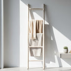 A tall, minimalist wooden ladder towel rack with three rungs, styled with three textured beige and cream towels, set against a white wall with dramatic sunlight and shadows.