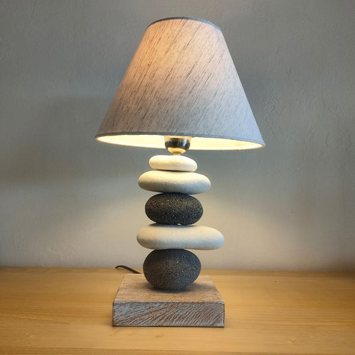 A bedside lamp from the Pebble Series, with a stack of pebbles serving as the base, placed on a wooden surface, with a grey lampshade, partially illuminated.