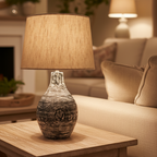 A wooden table lamp with a beige lampshade, turned on, providing a warm light. The lamp has a smooth, rounded base and a narrow, cylindrical shade.
